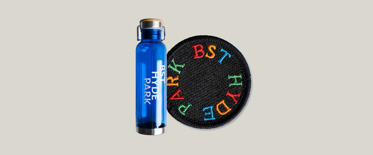 A blue glass water bottle labeled HYDE PARK stands upright next to a round black patch with colorful embroidered letters spelling BST HYDE PARK in a circle.